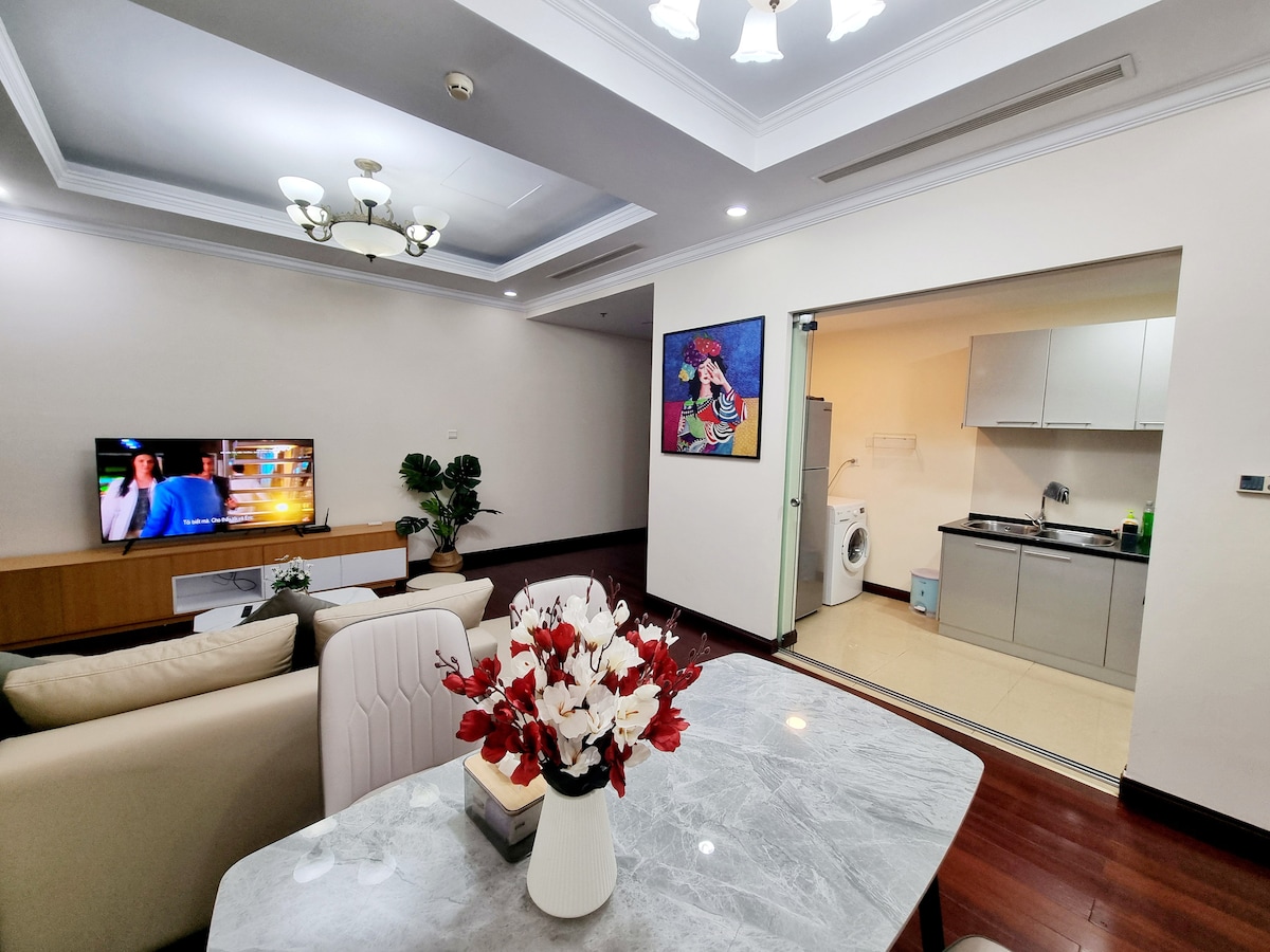 A spacious living area is shown, featuring a comfortable sofa and a coffee table adorned with decorative flowers. A wall-mounted Smart TV is visible, along with an open kitchen area equipped with modern appliances, including a washing machine. Natural light fills the space through bright overhead lighting.