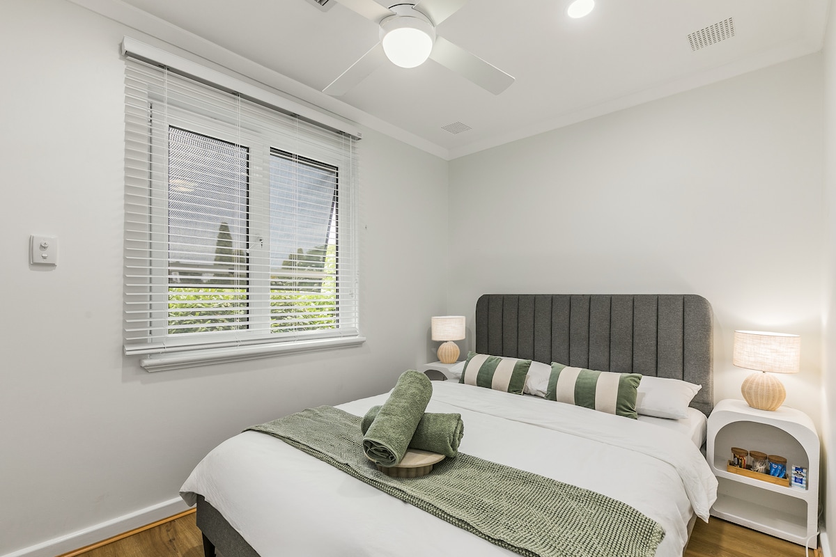 A light, airy bedroom features a spacious bed adorned with neatly arranged pillows and a textured throw. Soft, natural light filters through a large window dressed with blinds. A small bedside table holds a lamp, complementing the calming color palette of greens and whites.