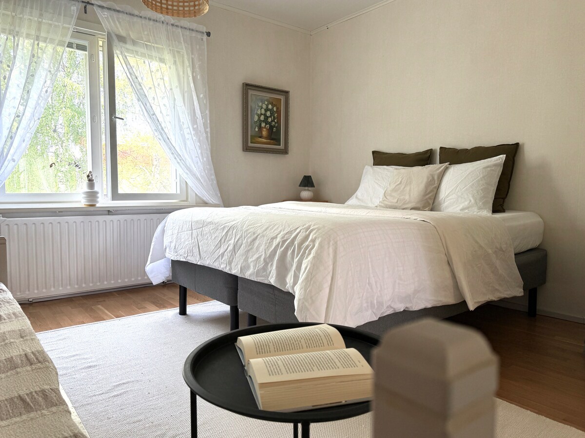 A spacious sleeping area features a large bed with a simple white bedspread and two accent pillows. Natural light filters through sheer curtains draping over a window, illuminating a small reading table with an open book. A neutral-colored rug lies beneath the bed.