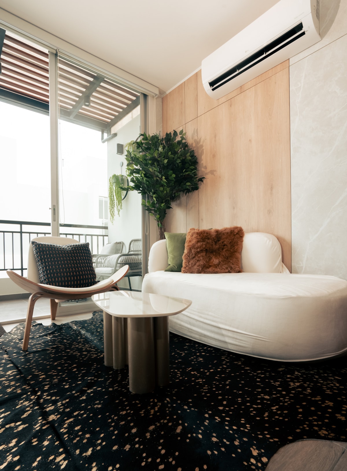 A modern living area is highlighted by a plush white sofa and a cozy chair. A small circular table sits at the center, while a large rug adds texture to the floor. Green plants add a touch of nature near expansive glass doors leading to a balcony.