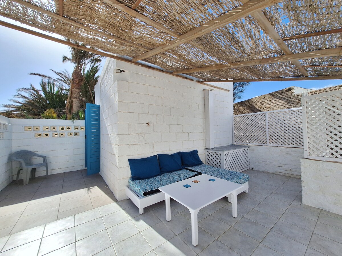 A relaxing outdoor space is furnished with a low table and a comfortable seating area, adorned with blue cushions. A shaded structure made of woven materials provides protection from the sun, surrounded by white walls and greenery in the background.