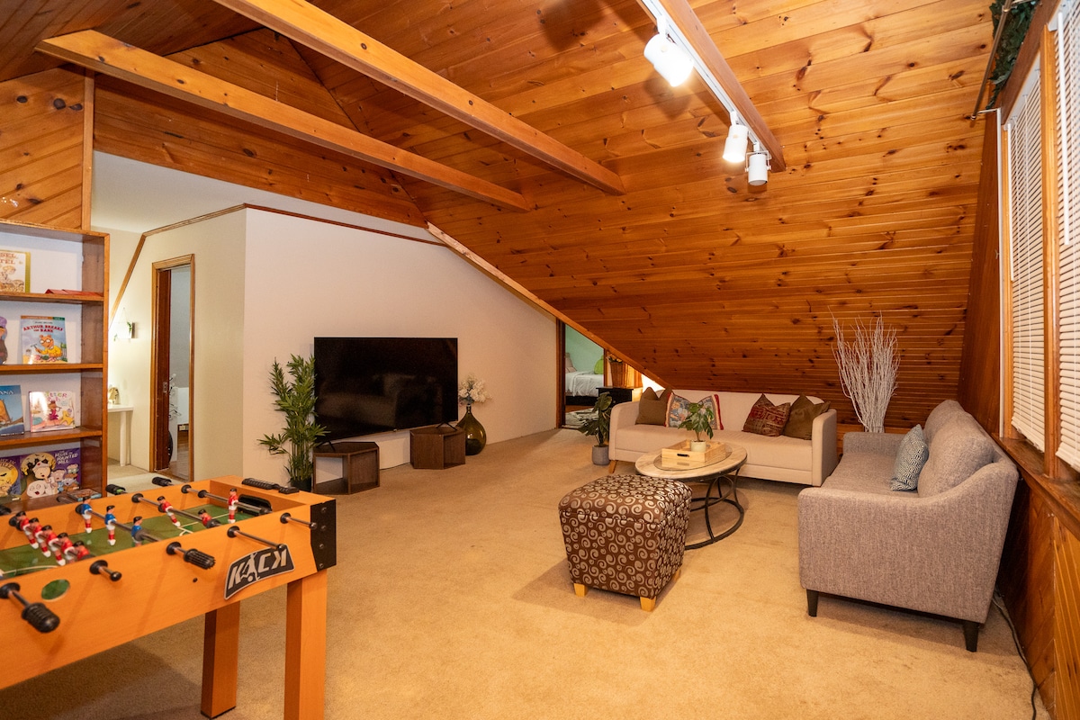 An attic common room showcases wooden beams and warm tones, featuring a cozy seating area with a light gray sofa and armchair. A 70-inch smart TV is mounted on the wall. A foosball table adds entertainment options, while shelves with books and games are visible.