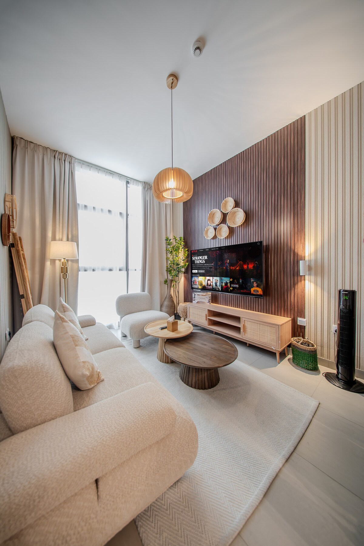 A modern living area features a comfortable light-colored sofa with plush cushions, positioned near a round coffee table. A television is mounted on the wall, and a stylish cabinet holds decorative items. Soft light filters through large windows, with textured curtains framing the view.