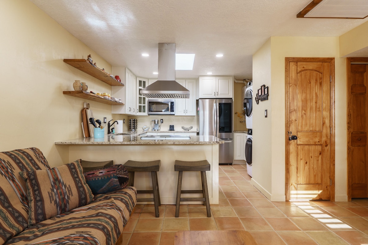 The kitchen features a central island with a stainless steel range hood above, surrounded by modern cabinetry and sleek countertops. Shelves display decorative items. A comfortable sofa with patterned upholstery is positioned nearby, complemented by tile flooring that adds warmth to the space.