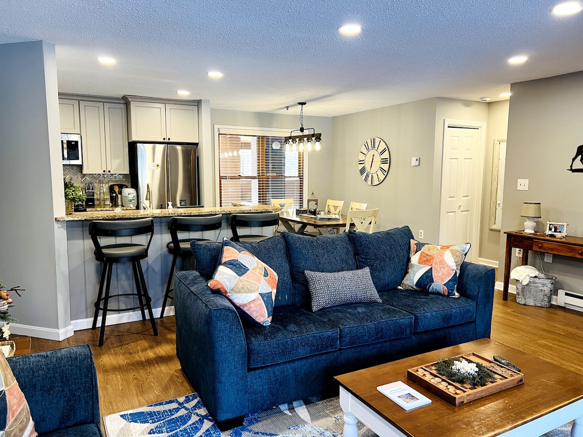 The living area features a spacious blue sofa adorned with decorative pillows, complemented by a wooden coffee table. An open kitchen with bar seating is visible in the background, along with a dining area illuminated by natural light from large windows.