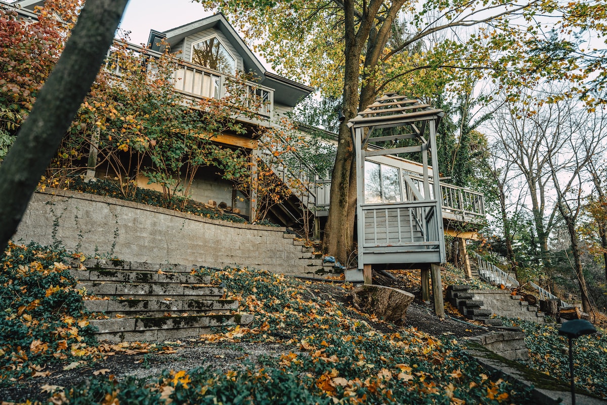 A serene outdoor view of the home's exterior, showcasing a multi-level structure surrounded by vibrant autumn foliage. Steps lead up to the entrance, with a decorative gazebo positioned prominently, offering an inviting space among the natural surroundings.