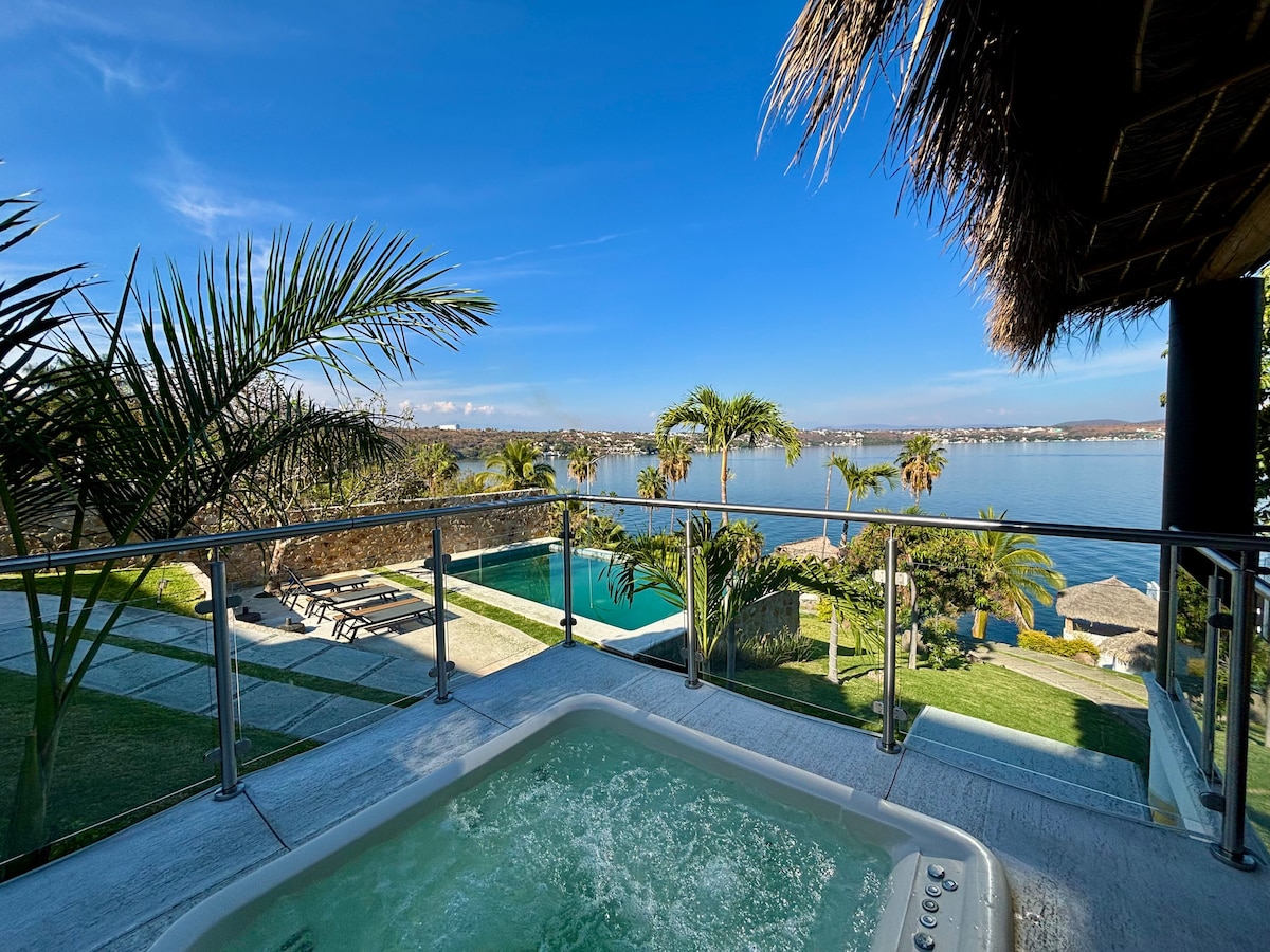 A hot tub is visible in the foreground, with a view of a serene lake and a well-maintained lawn. An infinity pool is situated beside the hot tub, palm trees are scattered throughout the landscape, and clear blue skies stretch above the tranquil setting.