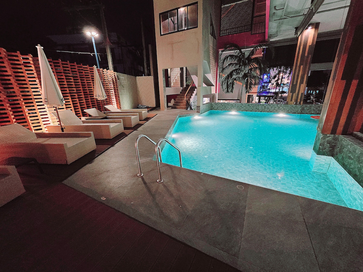 A serene pool area is featured under soft lighting, with clear turquoise water inviting a refreshing swim. White lounge chairs are positioned nearby, offering a relaxing space to unwind. The surrounding architecture includes a mix of modern design elements and lush greenery.