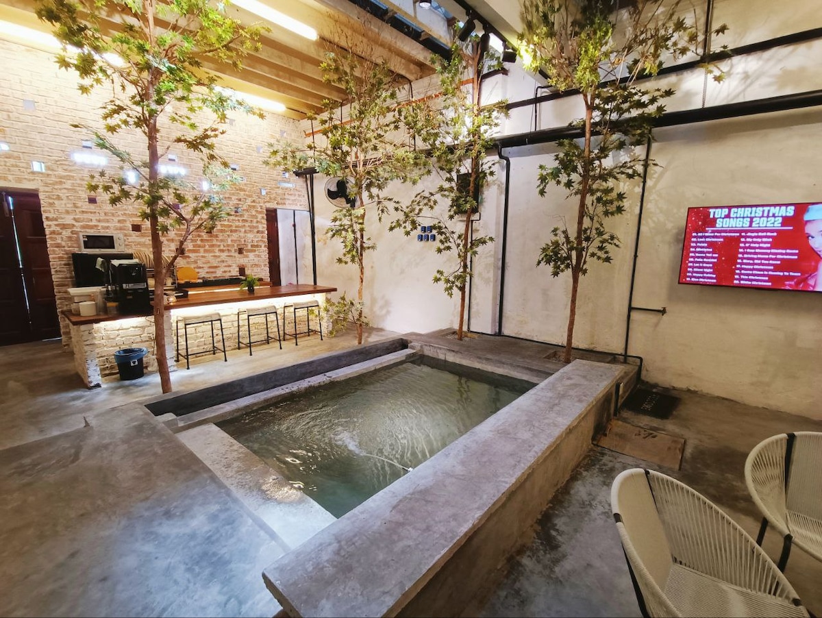 The private dip pool serves as a focal point of the space, surrounded by natural elements that enhance the industrial design. The kitchenette area is visible in the background, featuring bar-style seating and modern appliances. Soft lighting and plant accents contribute to a serene atmosphere.