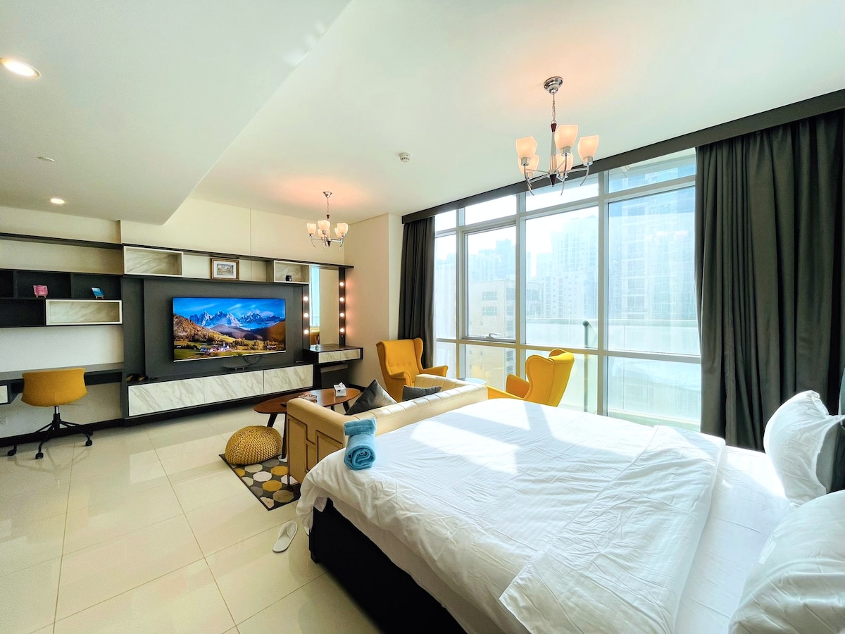 A spacious studio suite features a large window allowing natural light to fill the room. The seating area includes a modern sofa and two yellow accent chairs. A large television is mounted on the wall opposite the bed, creating an inviting space for relaxation.