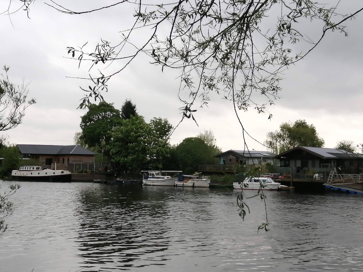 The image captures a serene waterscape featuring the River Trent. Several boats are docked along the riverbank, surrounded by lush greenery and residential structures. Overcast skies reflect softly on the water's surface, enhancing the calm atmosphere of the scene.