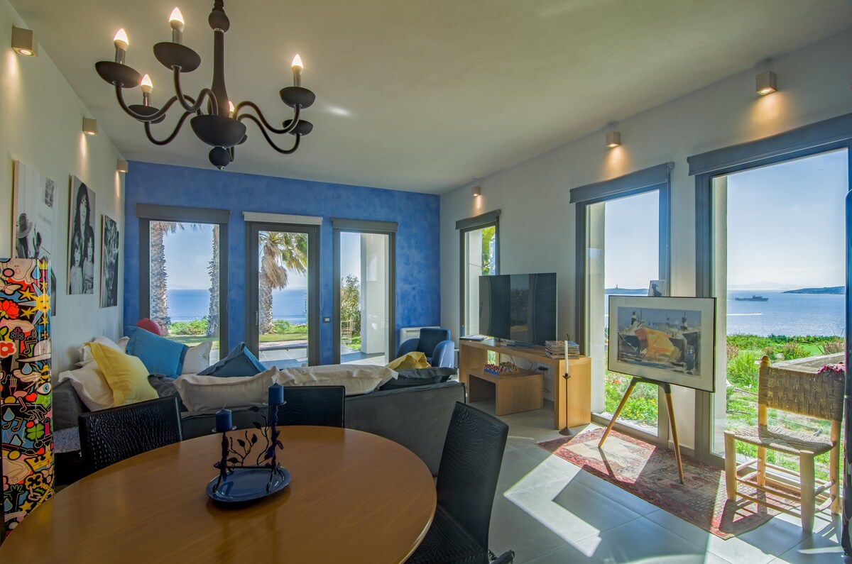 A spacious living area is highlighted by large windows, providing unobstructed sea views. Soft seating is arranged around a coffee table, while a dining table sits nearby. Natural light floods the space, enhanced by a modern chandelier. Decorative artwork adds character to the room.