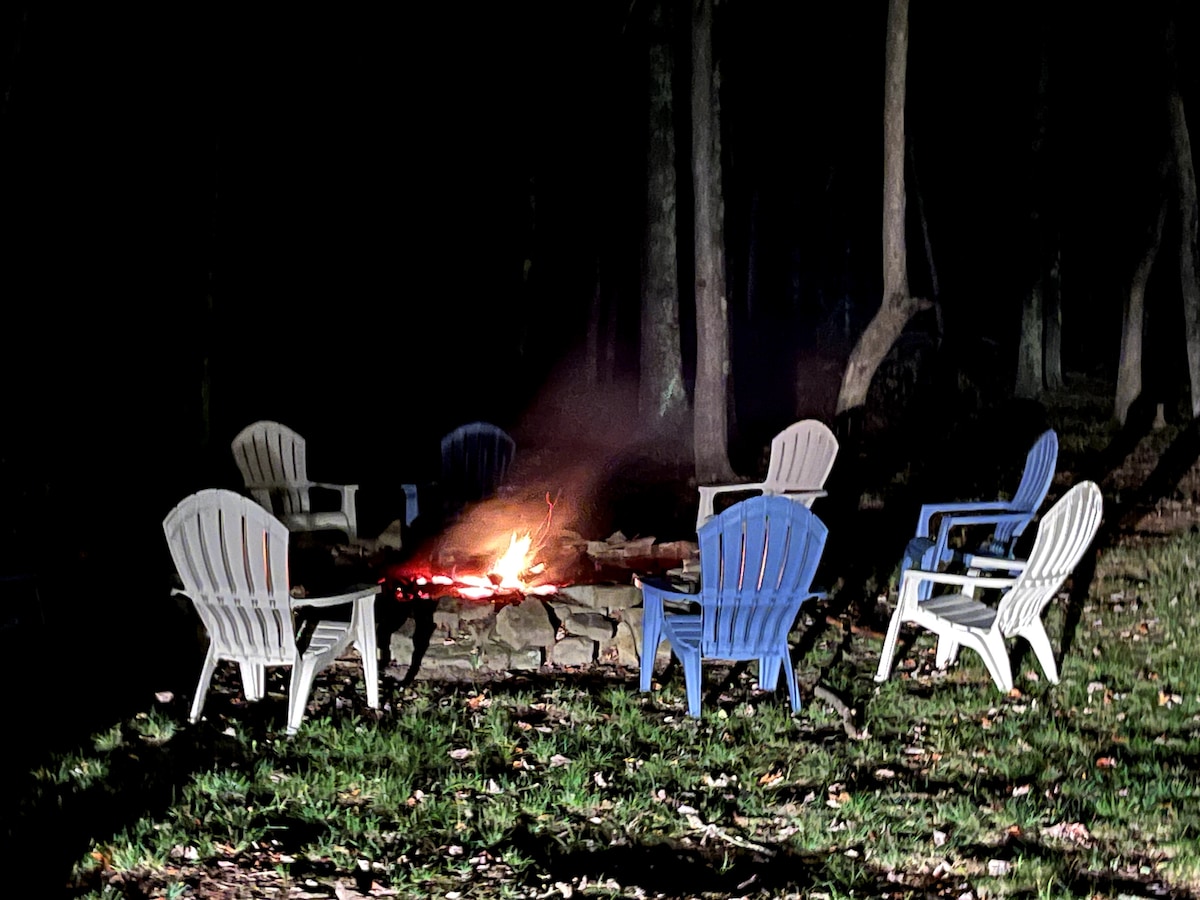 A cozy fire pit area is surrounded by several white and blue adirondack chairs, arranged to face the crackling fire. The setting is framed by tall trees, providing a sense of seclusion and nature. Soft grass covers the ground, creating a comfortable atmosphere for gatherings.