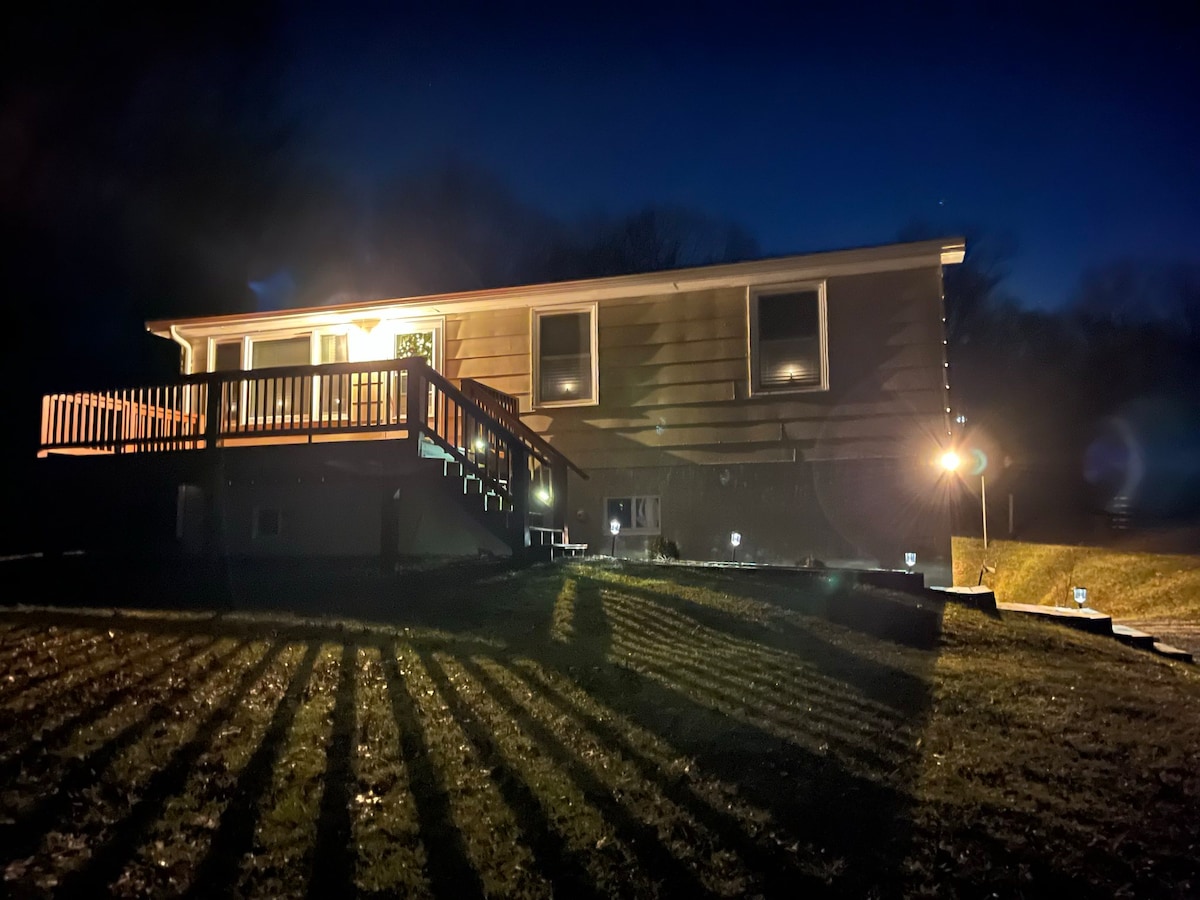 The cabin is illuminated at night, showcasing its welcoming exterior. A deck extends from the front, illuminated by soft lighting. Shadows are cast on the ground, highlighting the grassy area surrounding the cabin. The peaceful setting conveys a serene atmosphere.