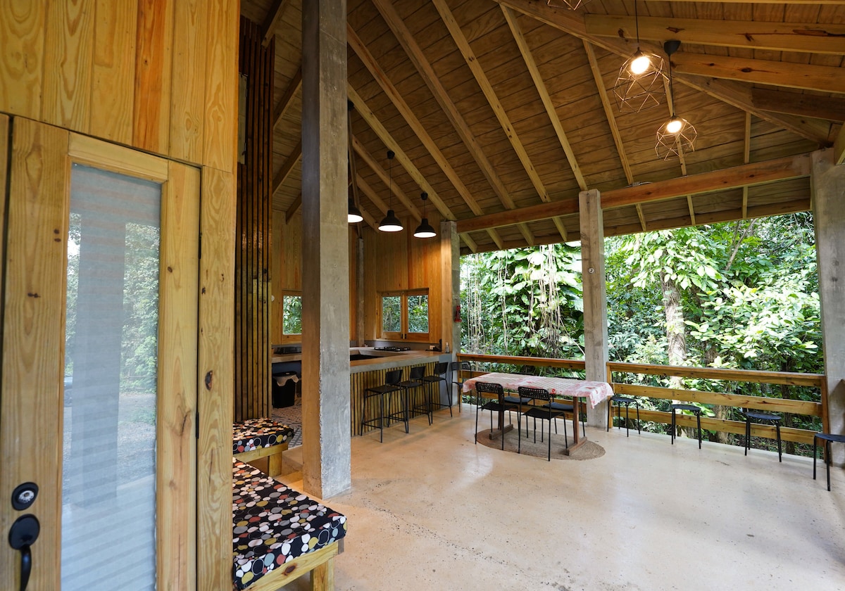 An open-plan interior features natural wood finishes and exposed beams. Large windows provide views of the surrounding forest. A dining table for six is positioned beside a kitchen counter, while cozy seating areas invite relaxation. Soft lighting enhances the spacious feel of the area.