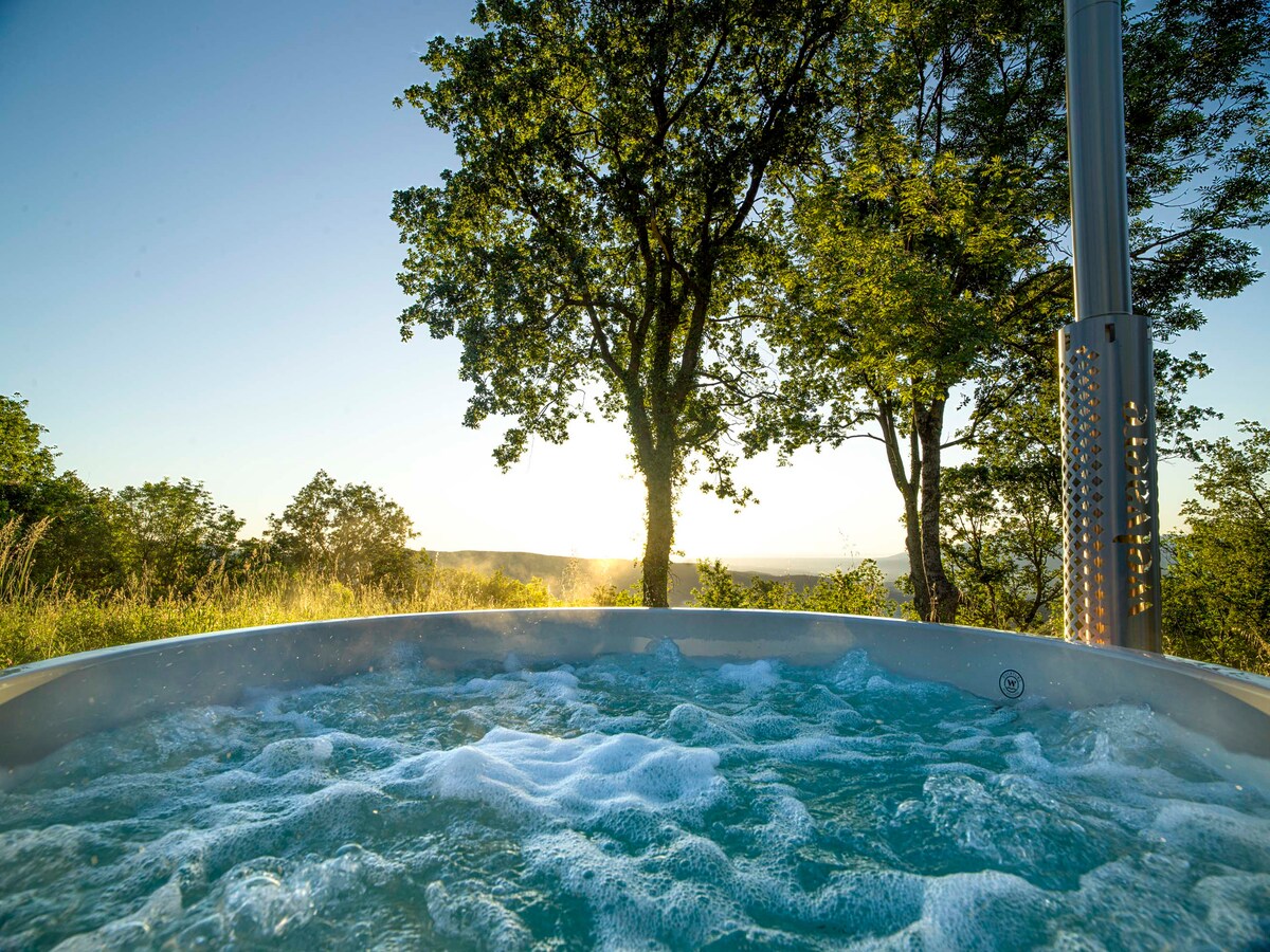 A wood-fired hot tub is filled with bubbling water, set in a natural landscape. The surrounding trees frame the scene as sunlight filters through, illuminating the vibrant greenery. A distant view of rolling hills is visible in the background, enhancing the tranquil atmosphere.