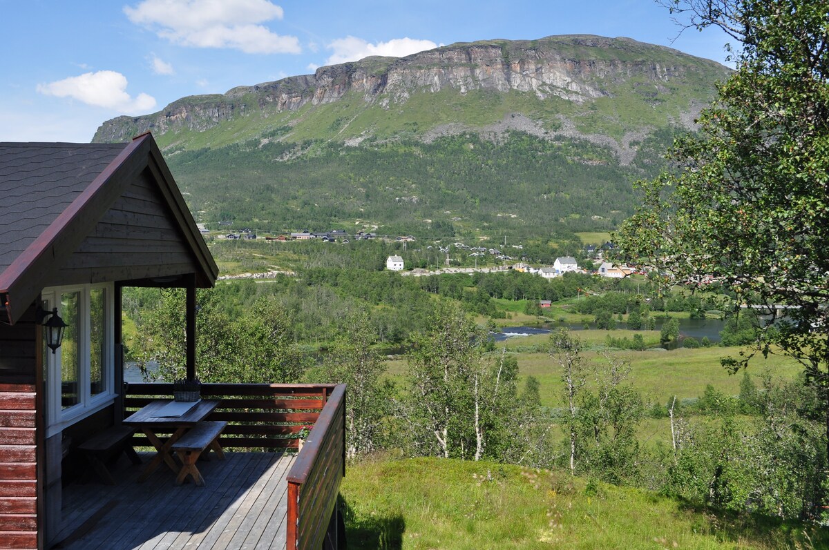 A wooden terrace is shown with a picnic table, overlooking a wide valley surrounded by greenery. In the distance, mountains rise against a clear blue sky, while a small town and a calm body of water are visible below.