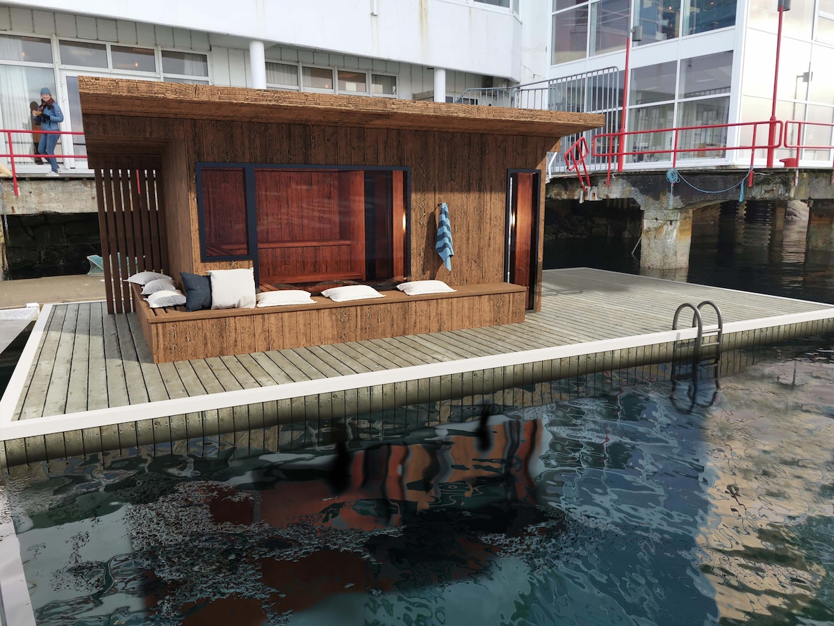 A wooden structure is situated on a floating platform above calm water. The design includes large windows and a shaded resting area with several cushions. A ladder leads to the water, providing easy access for swimming or relaxation.