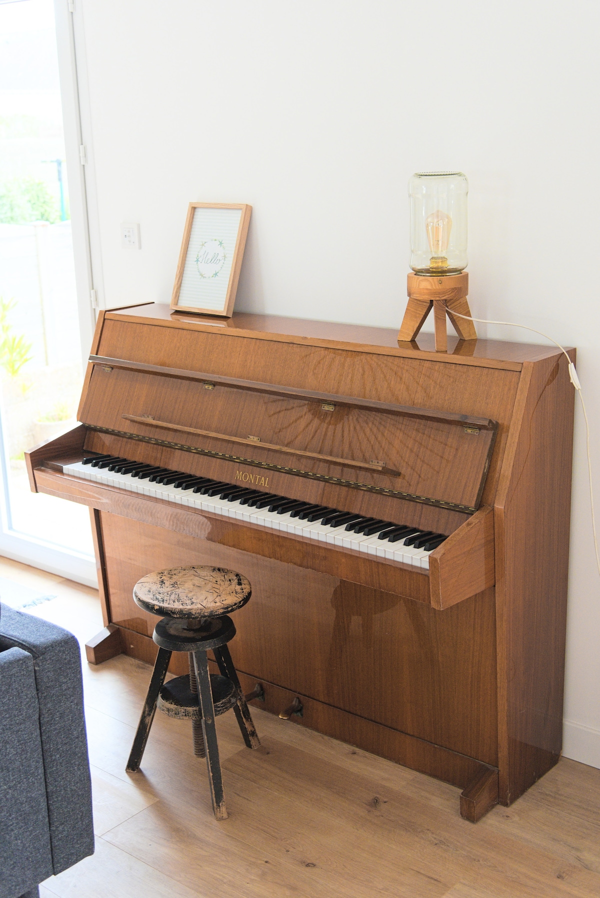A wooden piano stands against a light wall, featuring an array of keys and an elegant finish. A rustic stool is placed in front, accompanied by a stylish lamp on a small wooden stand nearby. Natural light enters through a window, enhancing the space.