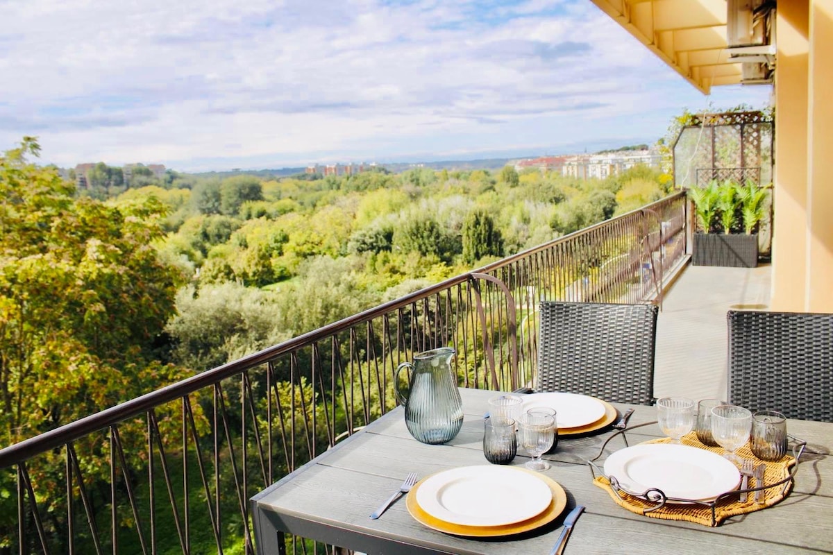An outdoor terrace is shown, featuring a dining table set with plates, glasses, and a water pitcher. Lush greenery and trees are visible beyond the railing, providing a serene view of the surrounding landscape. The space is adorned with decorative planters.