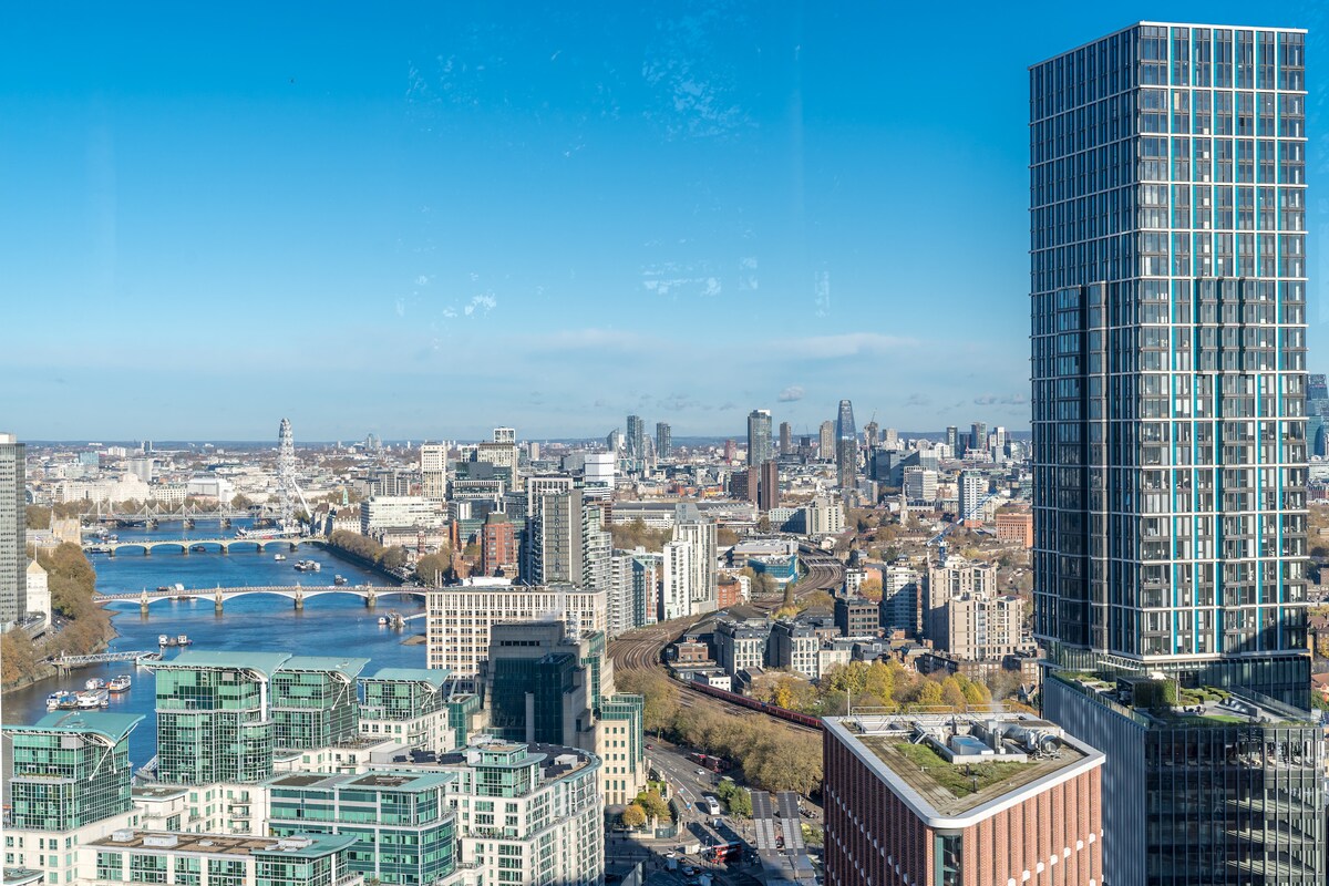 The image captures a panoramic view of London, showcasing the River Thames winding through the city. Modern skyscrapers line the skyline, with a prominent high-rise building in the foreground. A clear blue sky enhances the vibrant urban landscape.