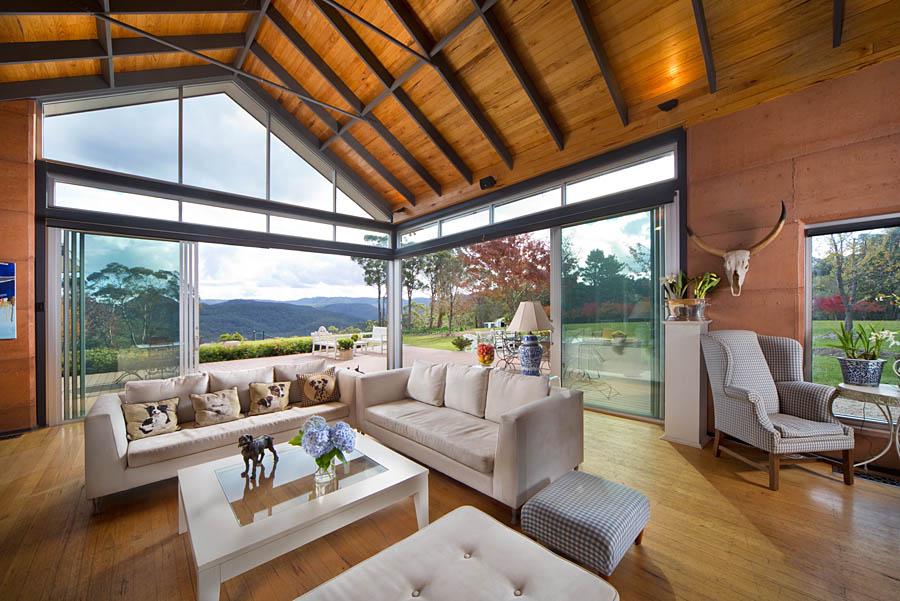 A spacious living area features large glass windows that provide sweeping views of the mountain landscape. Wooden beams highlight the vaulted ceiling. A comfortable sofa is arranged around a light coffee table, with a decorative lamp and floral centerpiece enhancing the serene ambiance.