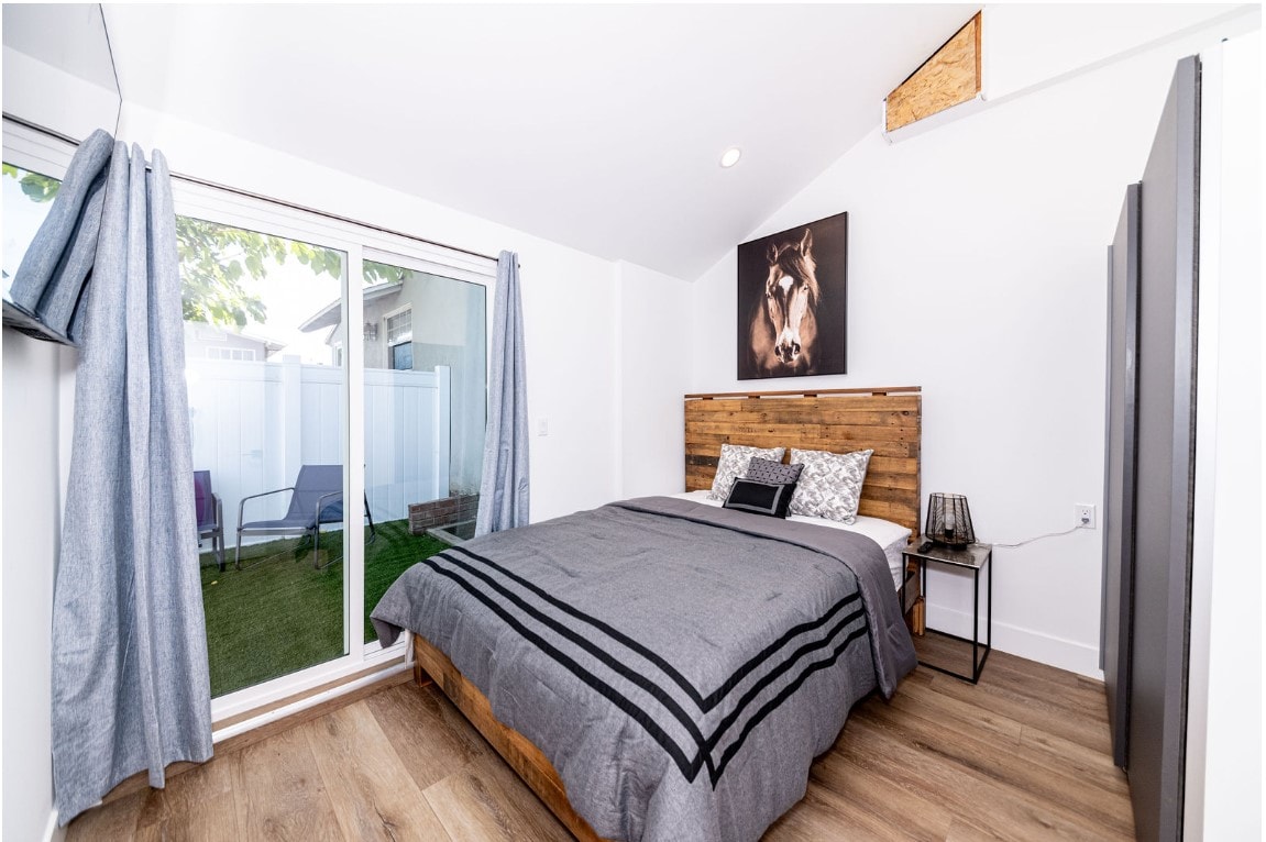 A spacious bedroom features a wooden bed frame, adorned with a simple gray coverlet and patterned pillows. Natural light floods in through large glass doors, leading to a grassy area outside. A piece of artwork depicting a horse is displayed above the headboard.
