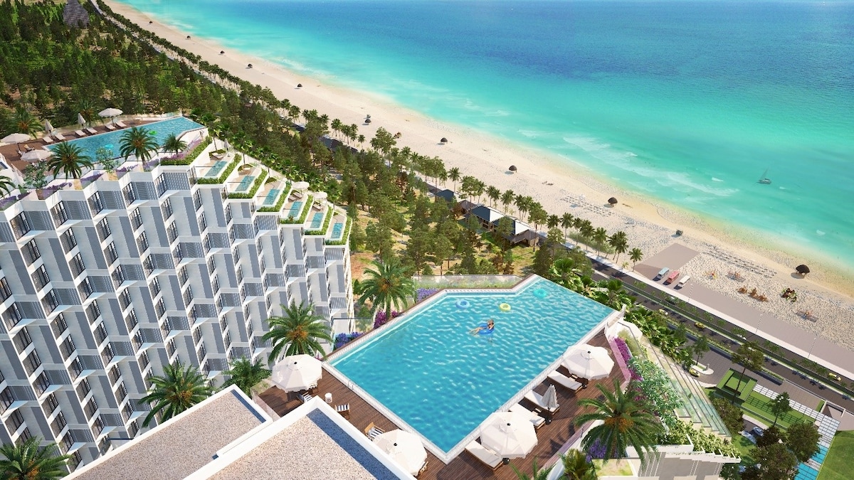 An aerial view of a resort features a large, inviting swimming pool surrounded by palm trees. The pool area is dotted with lounge chairs. The sandy beach and clear turquoise waters are visible in the distance, showcasing a serene coastal environment.