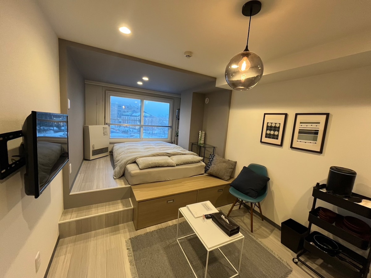 A compact bedroom features a raised sleeping area with a spacious bed topped with neutral bedding. Natural light fills the room through large windows, offering a view of the surroundings. A small table and a blue chair provide additional seating, complemented by a TV mounted on the wall.