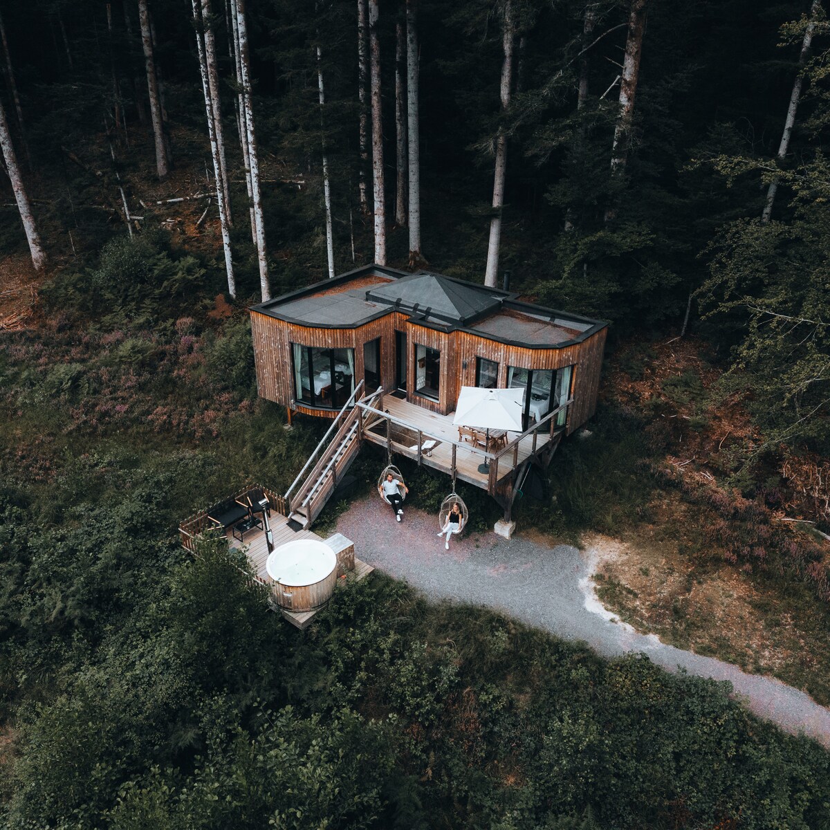 An intimate lodge nestled in the forest is comprised of large glass windows, allowing for a blend of indoor and outdoor spaces. A private hot tub is positioned on a wooden deck, while a stone pathway leads up to the entrance, flanked by lush greenery.