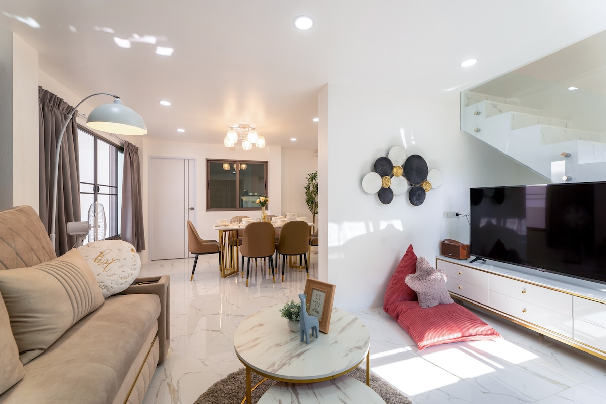 A modern living space is highlighted by soft, neutral-toned furnishings, including a plush sofa and cozy floor cushions. A round coffee table sits in front, while a large television complements the open area, which connects to a dining space decorated with stylish chairs and a contemporary chandelier.