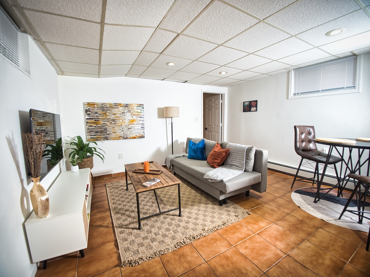 A spacious living area features a gray sectional sofa with vibrant orange and blue cushions. A wooden coffee table is centered on a patterned rug, with a floor lamp providing light. A dining table with metal legs is visible in the background, enhancing the room's utility.