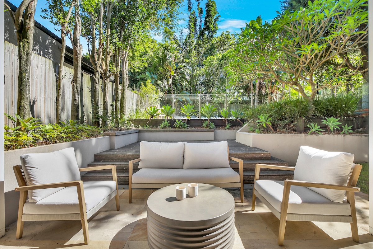 An inviting outdoor living area is presented with comfortable white seating arranged around a circular coffee table. Lush greenery surrounds the space, and steps lead up to tiered garden beds filled with various plants, creating a serene atmosphere.
