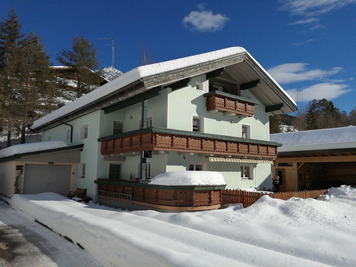 A three-story building features a light green exterior with wooden balconies adorned with decorative railings. A snowy landscape surrounds the home, with the roof blanketed in white snow. Clear blue skies and scattered clouds provide a serene backdrop.