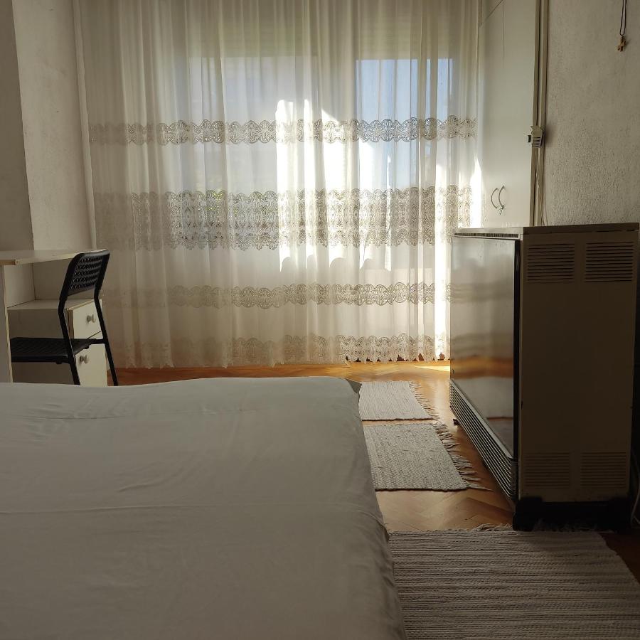 A bright bedroom features sheer lace curtains that filter natural light. A spacious bed is positioned near the window, complementing a simple desk and chair setup. A small heater is visible, enhancing comfort during cooler months.