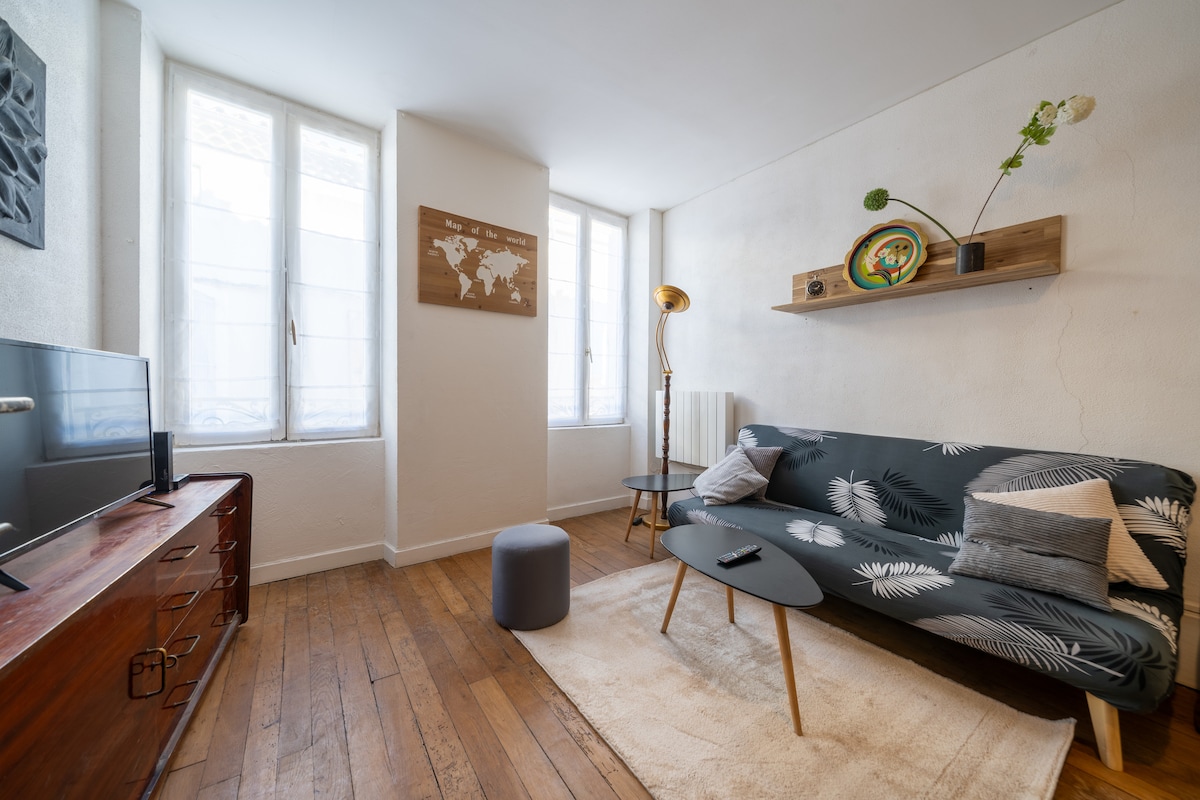 A inviting living area features a dark patterned sofa adorned with decorative cushions. A low coffee table sits in front, accompanied by a small stool. Natural light floods in through two tall, partially covered windows, illuminating wooden flooring and enhancing the room's modern decor.