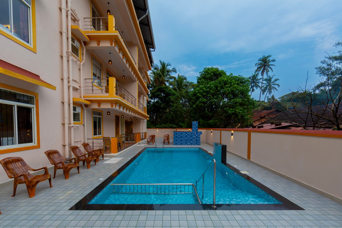 A clear view of a swimming pool surrounded by tiled flooring is presented. Several wooden lounge chairs are positioned near the pool. The property includes two multi-story buildings with balconies overlooking the pool area, complemented by lush greenery and palm trees in the background.