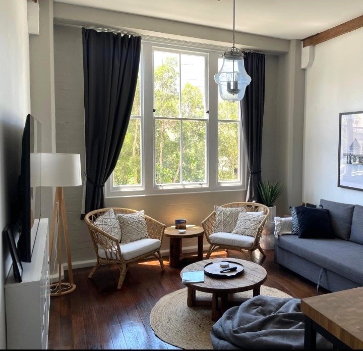 A bright living area is illuminated by natural light streaming through large windows adorned with dark curtains. Two wicker chairs and a round coffee table are arranged on wooden flooring, complemented by a cozy rug. A sofa and a TV are positioned along the opposite wall.