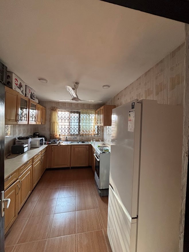 A functional kitchen is featured, with wooden cabinetry and a white refrigerator. Appliances, including a microwave and oven, are present. Natural light filters through a window, illuminating the tiled floor. The layout provides ample space for movement and cooking activities.
