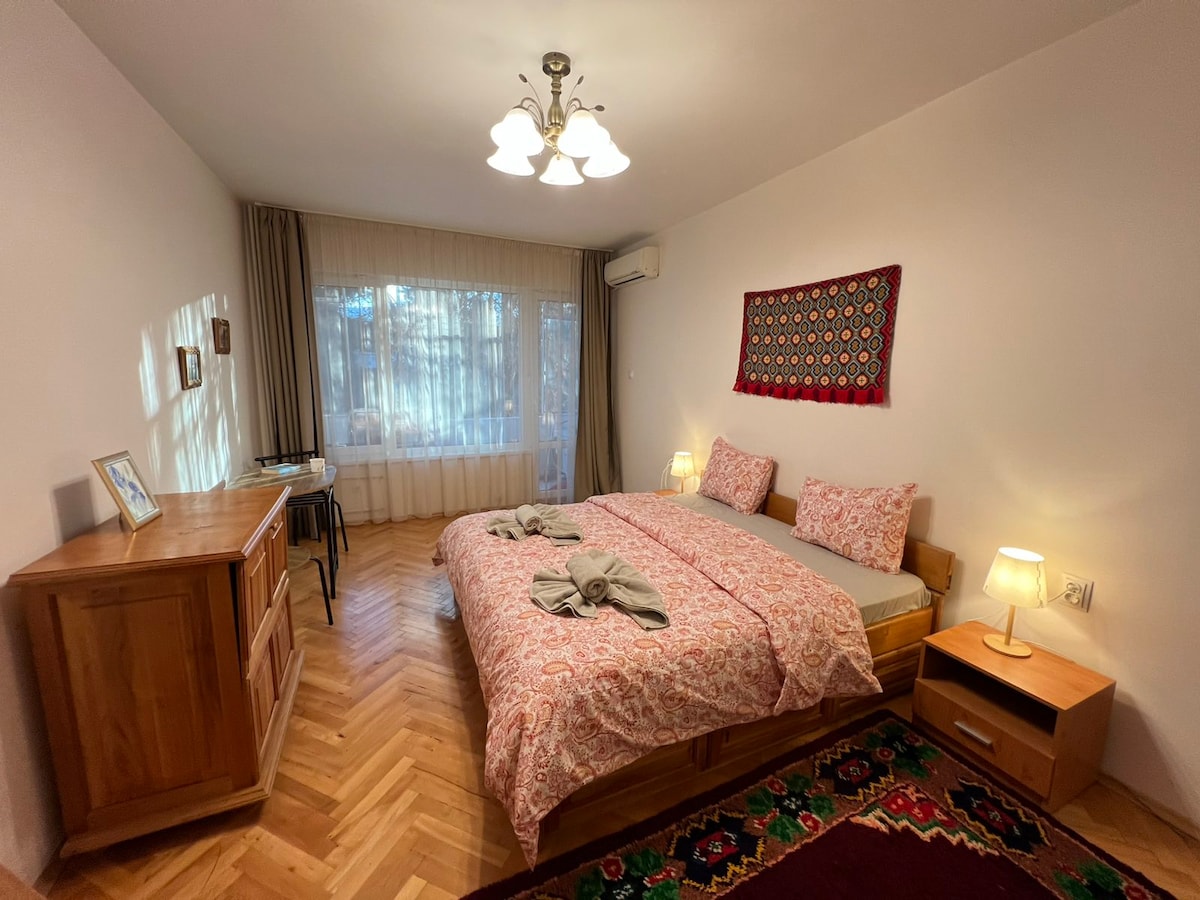 A warm and inviting bedroom features two spacious beds with patterned bedding, complemented by bedside lamps. A large window allows natural light to fill the room, while a traditional wall hanging adds character. A wooden dresser and desk are positioned, providing ample storage and workspace.