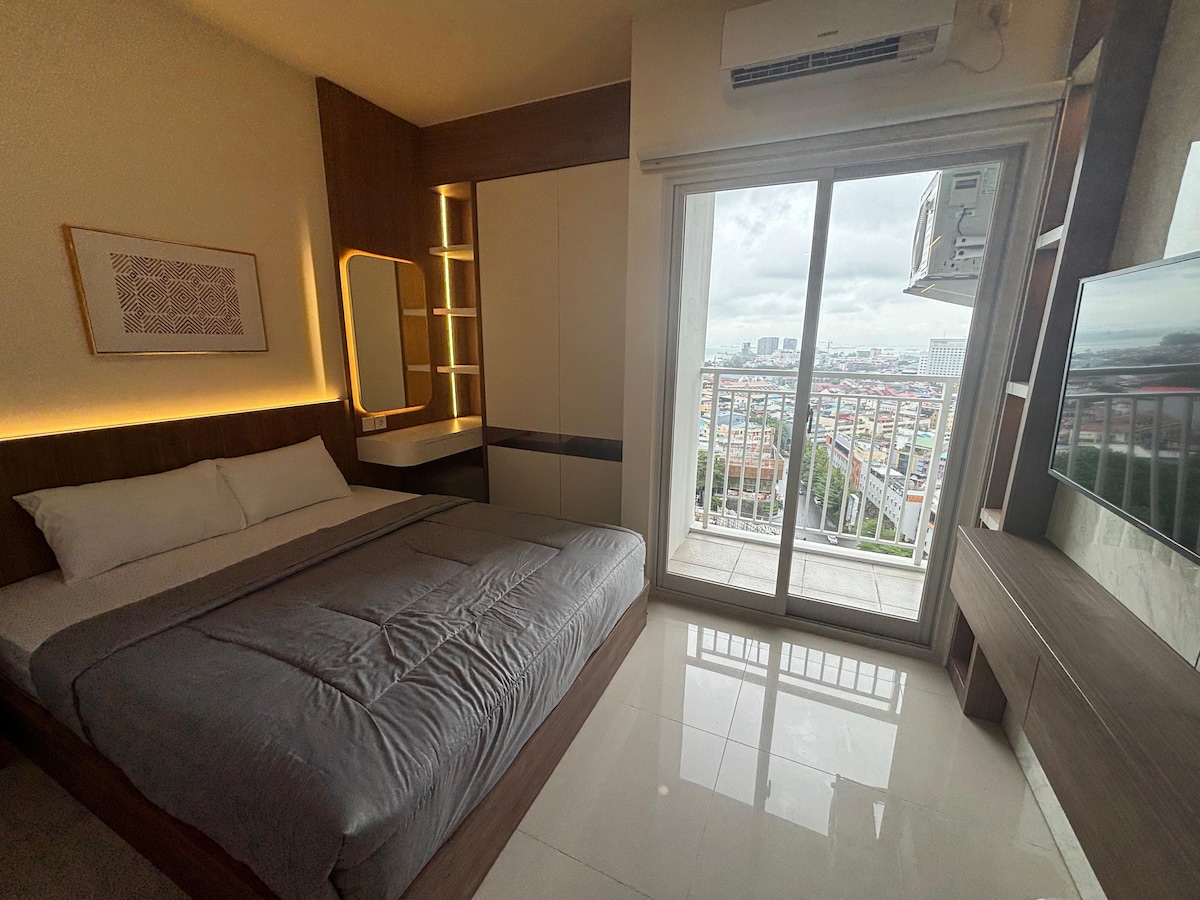 A well-lit bedroom featuring a spacious bed with gray bedding. A large window offers a view of the city, and a balcony door allows access to outdoor space. Minimalist cabinetry complements the modern decor, and a wall-mounted television adds convenience.