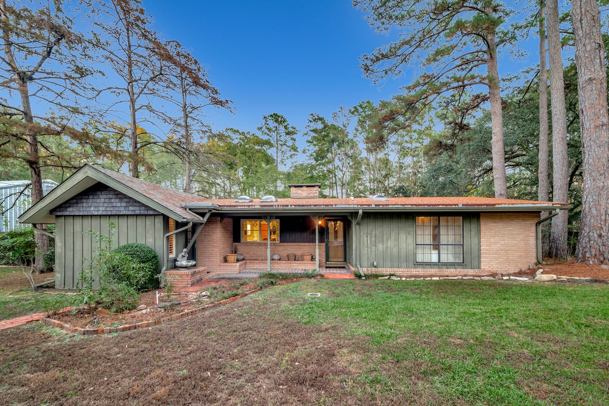 A charming cottage is framed by tall trees, showcasing a warm exterior of brick and wood. The inviting front porch features seating, with windows allowing light to illuminate the cozy interior. Lush greenery surrounds the well-maintained yard, enhancing the tranquil setting.