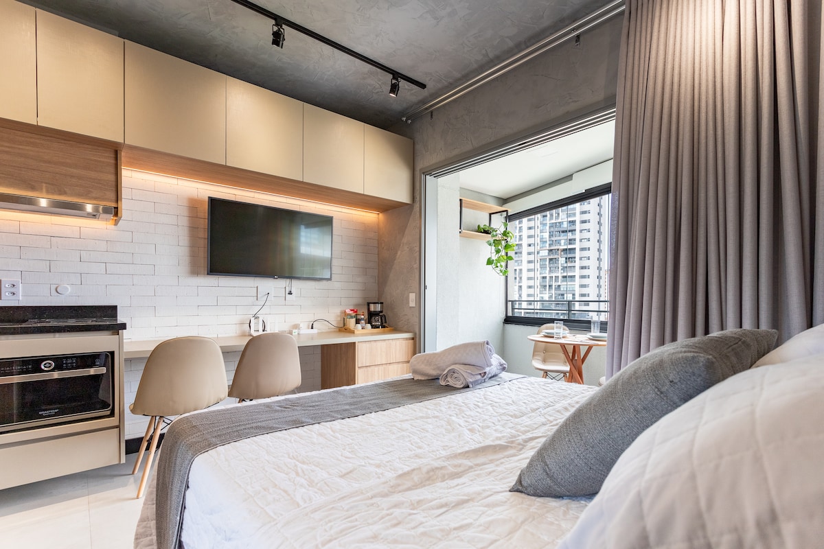 A modern studio space features a spacious bed with soft linens and a decorative throw. A kitchenette with stylish cabinetry occupies one wall, while a wall-mounted TV enhances the area. Natural light floods in from a large window with a small dining table on the balcony.