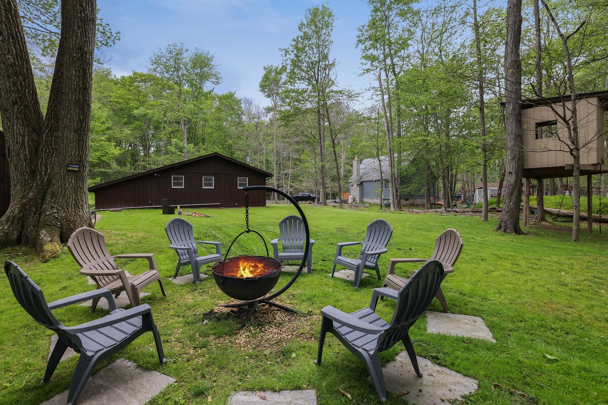 An inviting outdoor space showcases a circular fire pit surrounded by several adirondack chairs on a grassy area. Lush trees provide shade, and a treehouse can be seen in the background, creating a peaceful, natural setting.