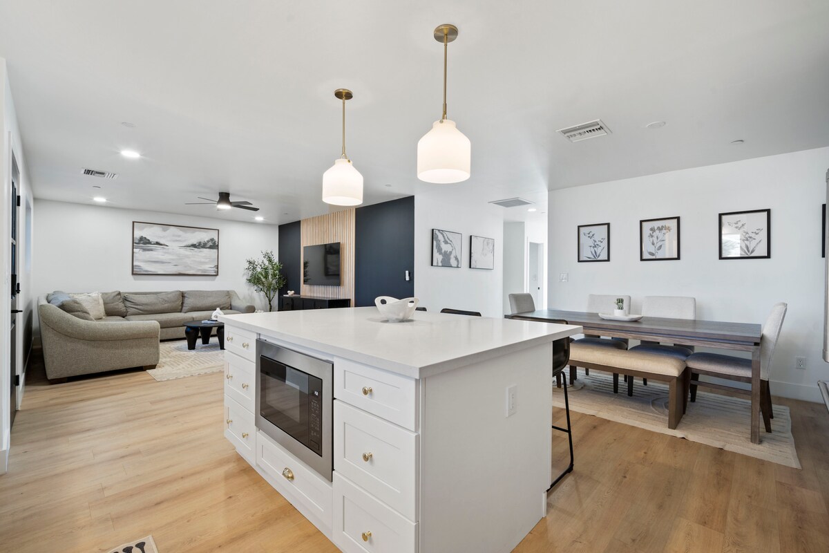 A modern kitchen showcases a central island with a white countertop and built-in microwave. Soft lighting is provided by pendant fixtures above the island. The adjacent dining area features a long table surrounded by chairs, while a comfortable seating area is visible in the background.