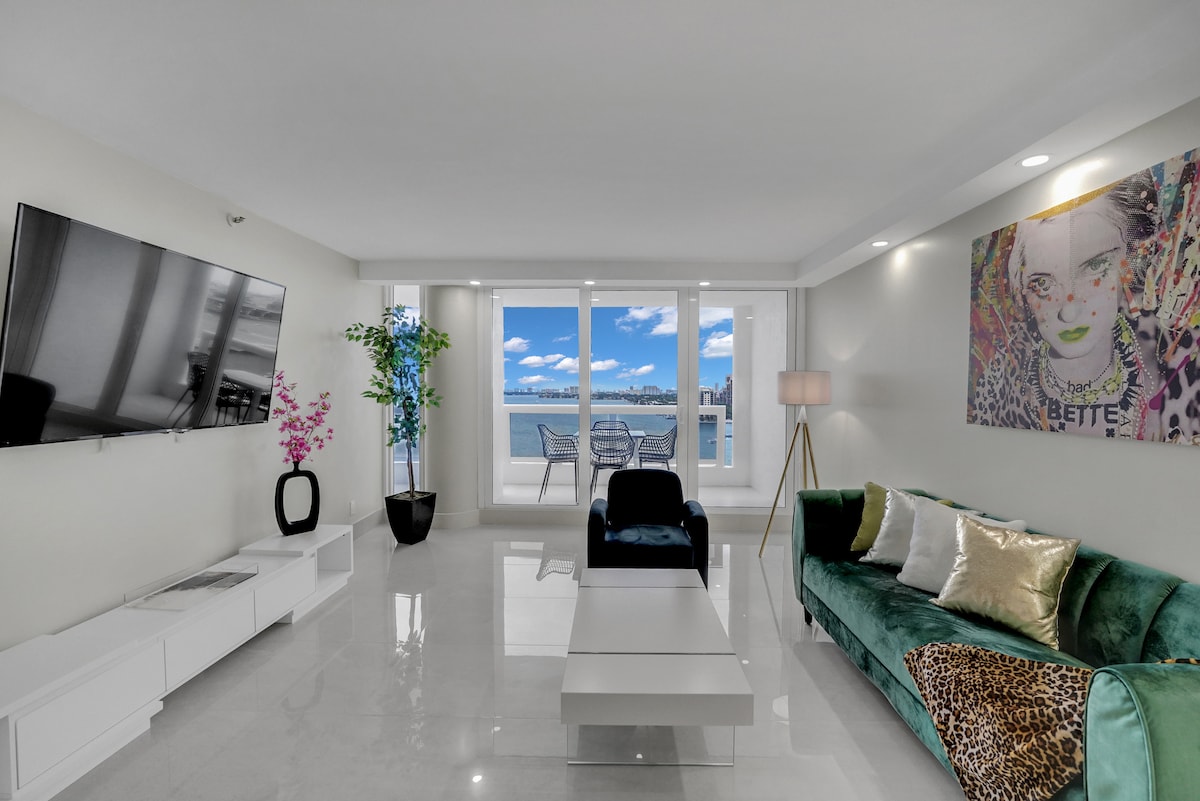 A modern living area is highlighted by a plush green sofa adorned with decorative pillows. A large wall-mounted TV is positioned across from a stylish coffee table. Floor-to-ceiling windows showcase stunning water views and access to the private balcony, enhancing the space's bright ambiance.