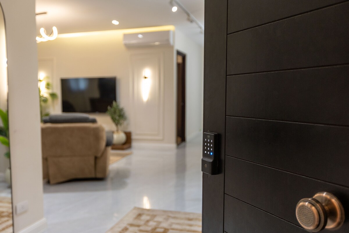A view from the entrance shows a modern living area featuring a large flat-screen TV mounted on the wall. A plush sofa is positioned nearby, with indoor plants adding a touch of greenery. Soft lighting creates a welcoming environment.