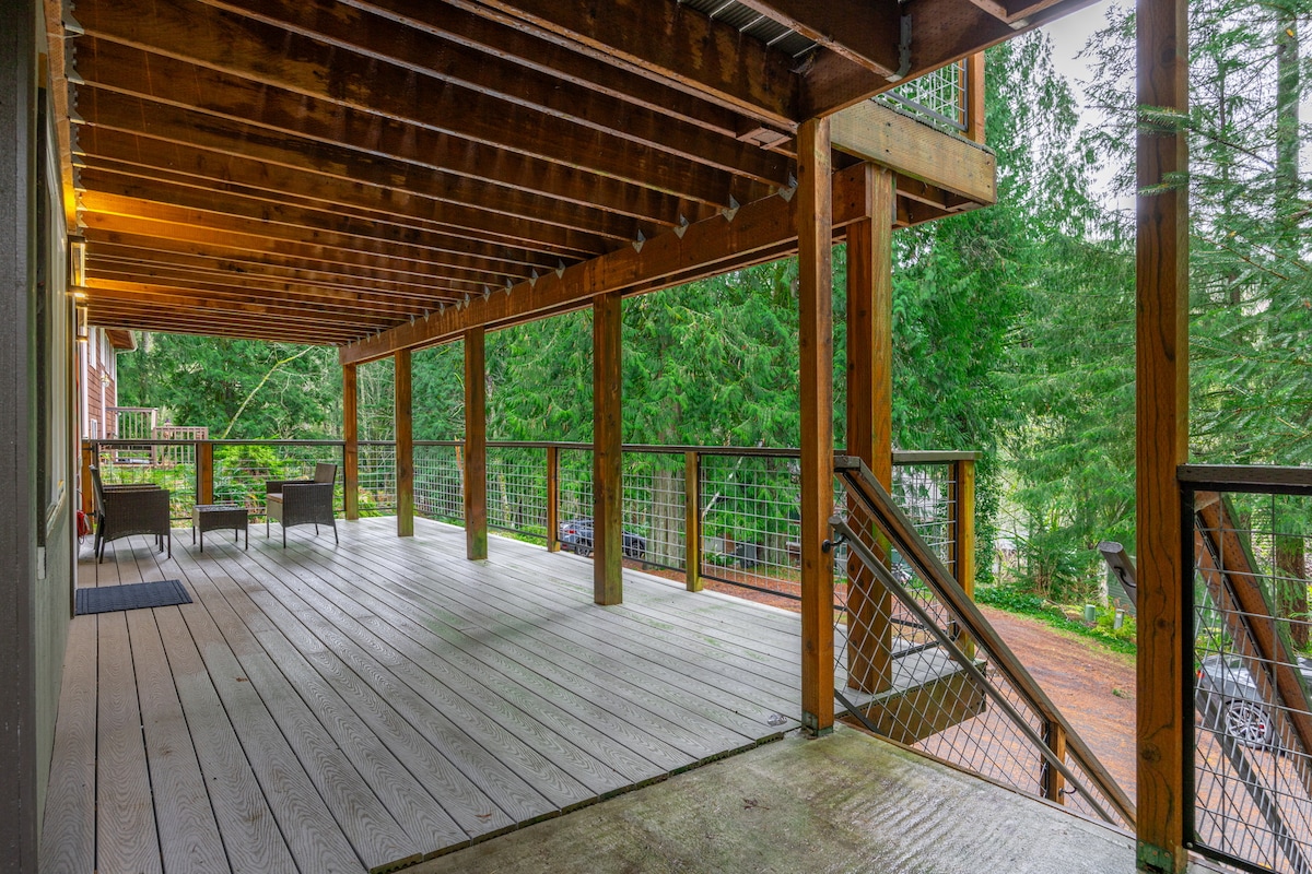 A spacious wooden deck is surrounded by lush greenery, offering a peaceful outdoor setting. Multiple seating options are visible, providing comfort and relaxation. A sturdy railing enhances safety while maintaining an unobstructed view of the forested area, inviting the experience of nature.