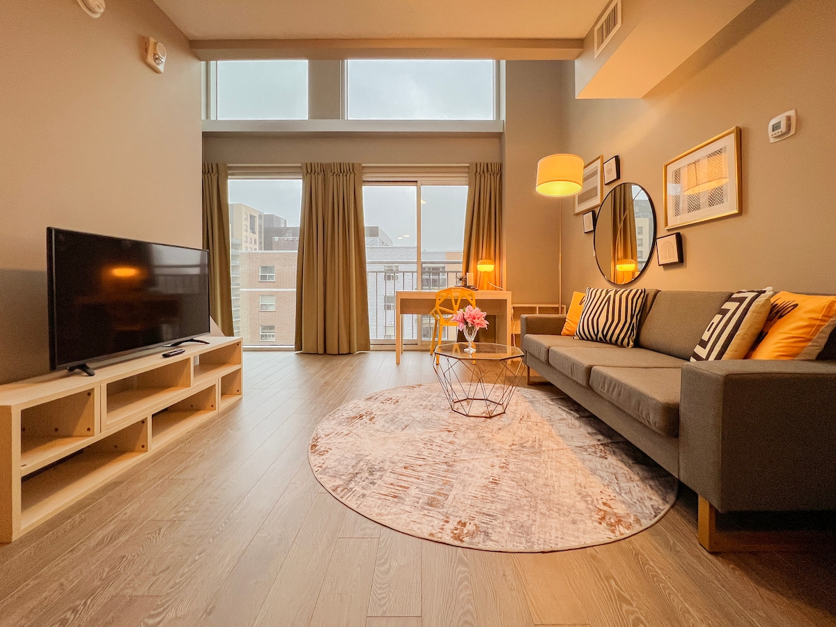 A spacious living area features large windows that provide ample natural light. A comfortable gray sofa is complemented by vibrant accent pillows. A modern circular rug lies under a stylish coffee table. A Smart TV is placed on a light wooden media console, with a dining table visible in the background.