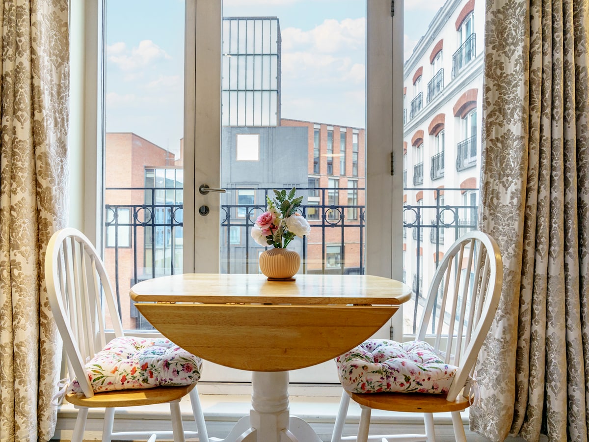 A small round table is set by a large glass door, allowing natural light to fill the space. Two white chairs with floral cushions are positioned around the table. A vase with flowers is placed at the center, offering a simple decorative touch.